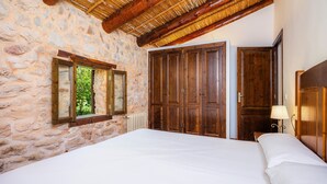 A bedroom with stone walls, a wooden ceiling, a large bed, and a window with a view of trees. - Apartment 'Es Sestador' with Mountain Views, Shared Pool and Wi-Fi (Illes Balears)