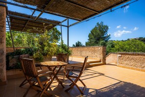 Une terrasse couverte, dotée d’une table et de chaises, entourée de plantes et offrant une vue sur le paysage.