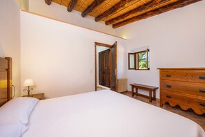 A bedroom with a bed, wooden dresser, nightstand, lamp, and a door leading to another room.