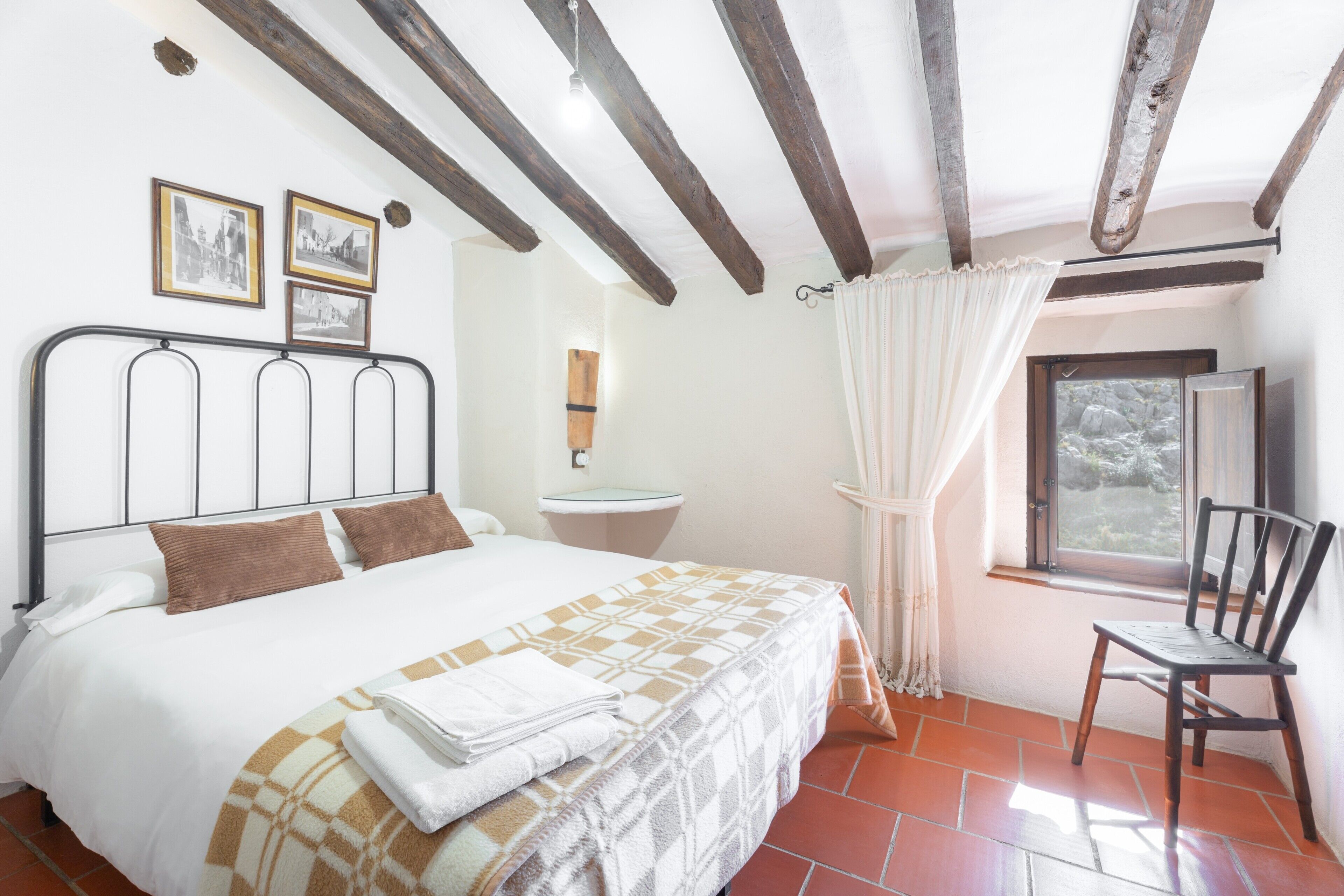 Ein Schlafzimmer mit Bett, Stuhl, einem Fenster mit Vorhängen und einem Wandbild.