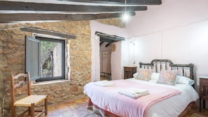 Un dormitorio con pared de piedra, cama de madera con almohadas estampadas, una silla de madera y una ventana con vistas.