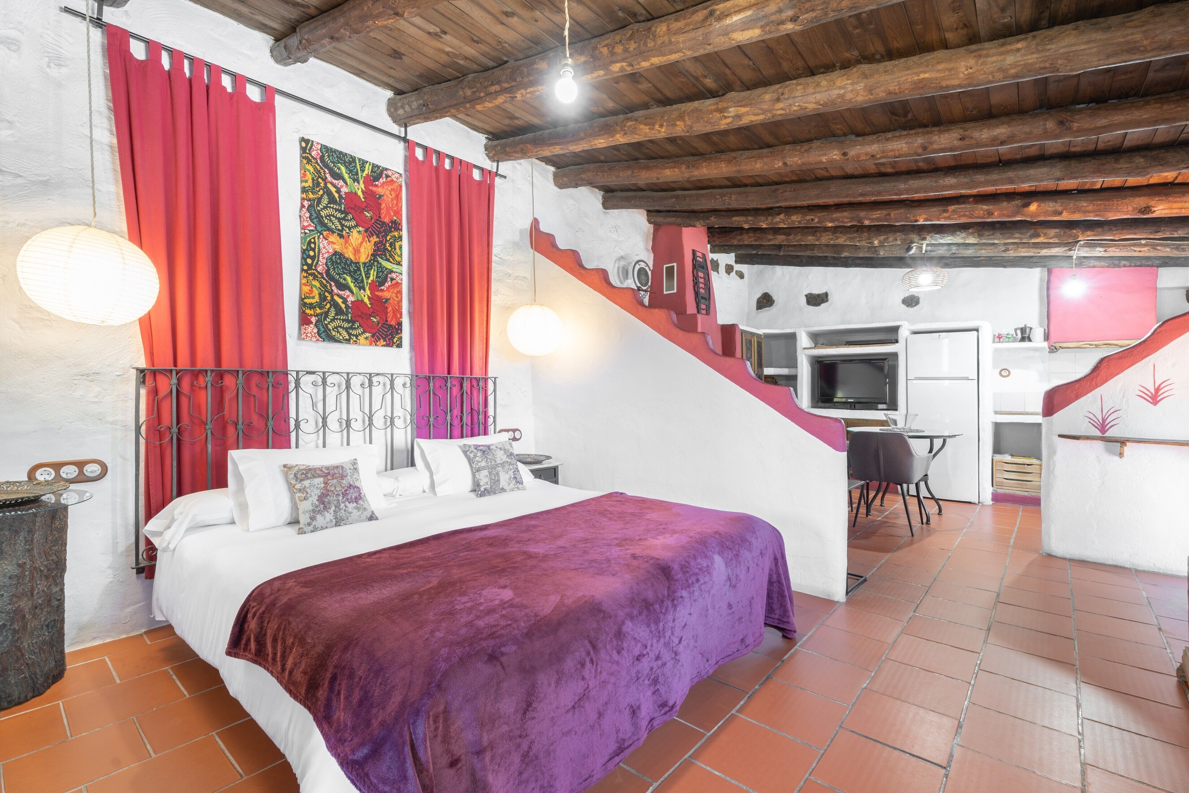 Une chambre avec un lit, des rideaux rouges, un escalier et un coin cuisine.