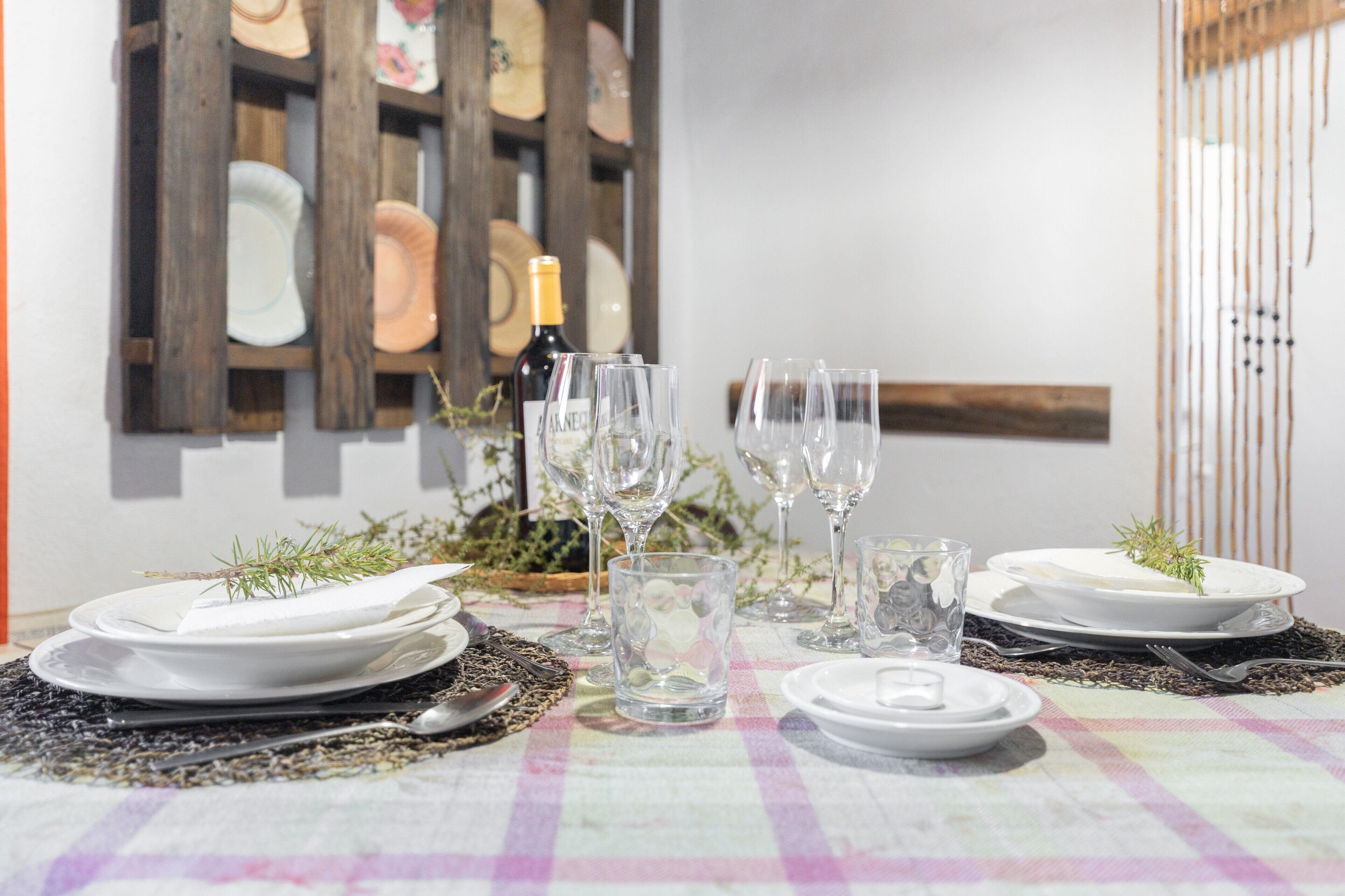 Una mesa dispuesta con platos, vasos y una botella, con mantel a cuadros y un fondo decorado con adornos de madera.