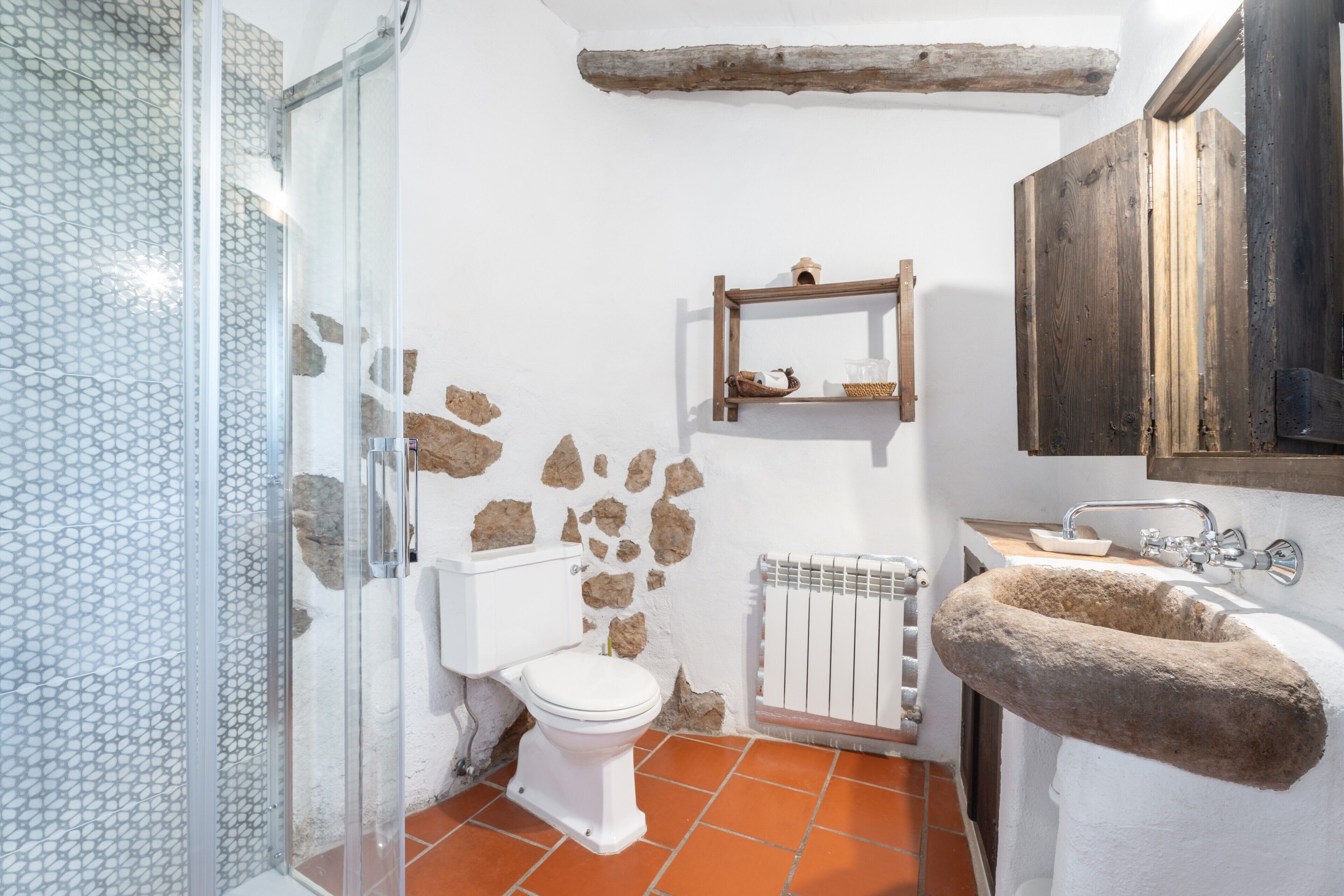 Un baño con cabina de ducha de vidrio, un inodoro blanco, un lavamanos de piedra y un estante de madera rústica.