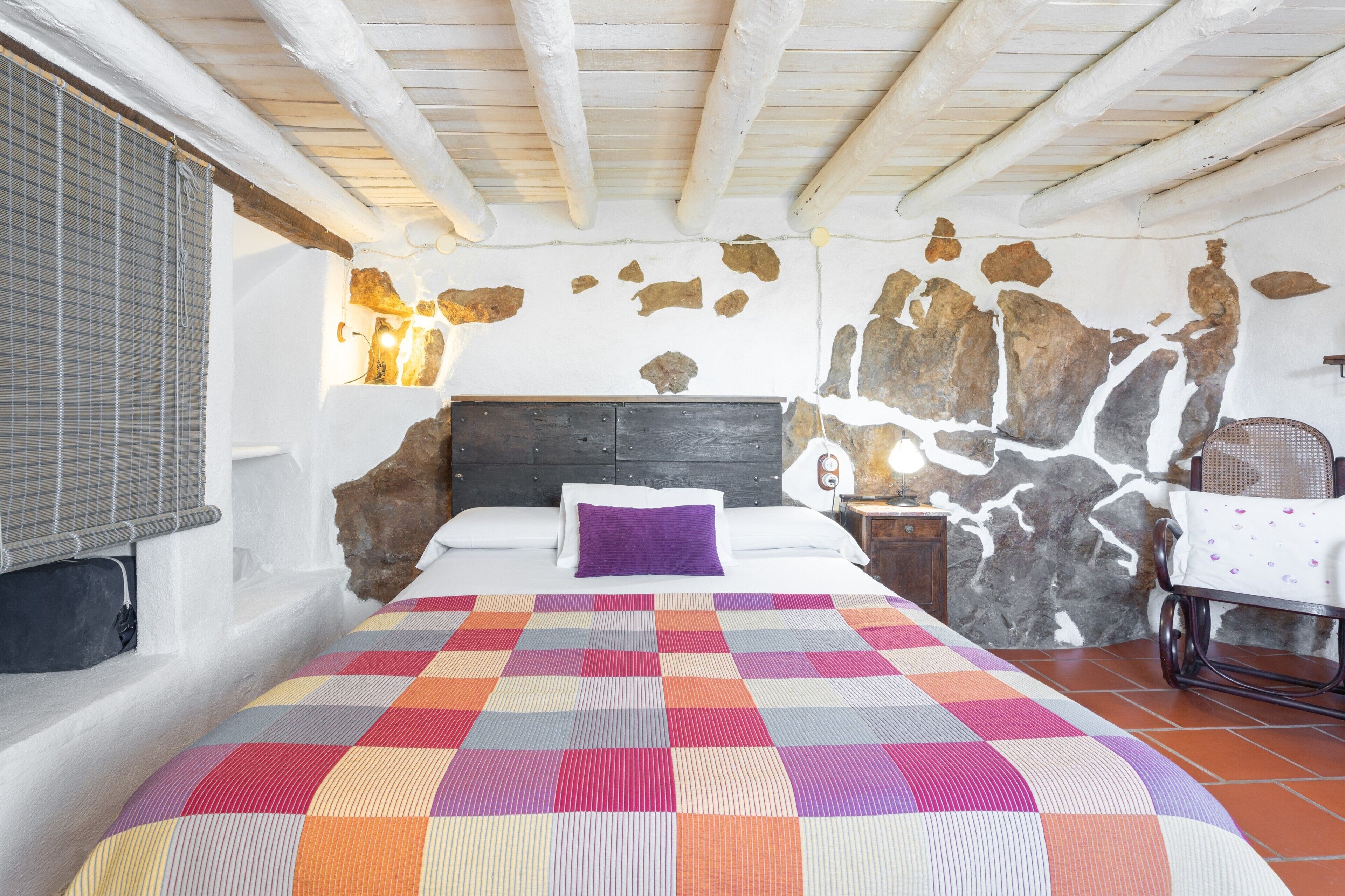 Ein Schlafzimmer mit einem Bett, einer farbenfrohen Steudecke, einer Steinwand, einer Holzbalkendecke und einem Stuhl.