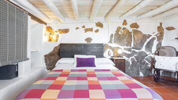 Ein Schlafzimmer mit einem Bett, einer farbenfrohen Steudecke, einer Steinwand, einer Holzbalkendecke und einem Stuhl.