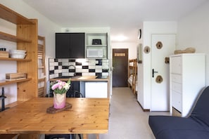 Cocina compacta con microondas, refrigerador y estantes de madera. Hay una mesa de comedor con un jarrón de flores. Se ve un armario blanco y un sofá en la habitación.