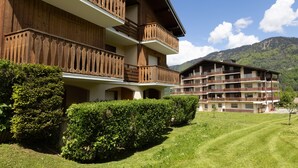 Un immeuble à plusieurs étages doté de balcons, entouré de pelouses et de haies bien entretenues, avec en toile de fond un paysage montagneux.