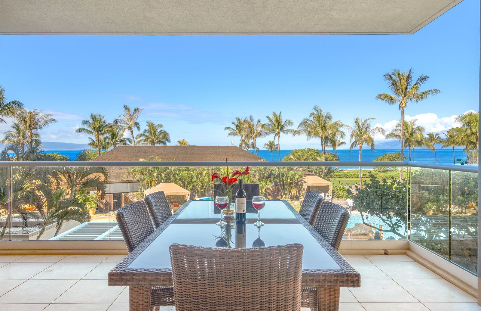Ein Balkon mit Glastisch und Korbstühlen, Blick auf einen Pool und Palmen mit Meerblick.