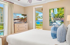 Habitación de hotel con una cama grande, un aparador, un cuadro de una escena tropical y vista al océano a través de una ventana.