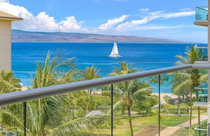 A balcony with a view of the ocean, palm trees, and a distant sailboat.