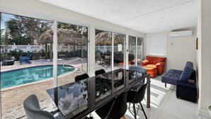 Un espace intérieur moderne doté d’un mur de verre donnant sur une piscine, d’un coin repas avec une table en verre et d’un espace détente avec des canapés orange et bleus.