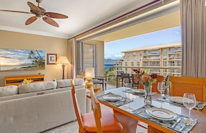 A hotel room with a balcony, a dining table set for two, a sofa, a ceiling fan, and a painting of a beach scene.