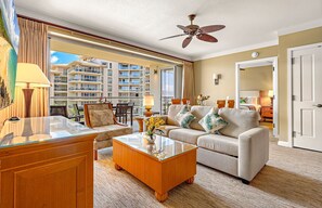A hotel room with a balcony, a ceiling fan, a sofa, a coffee table, and a view of the hotel buildings.