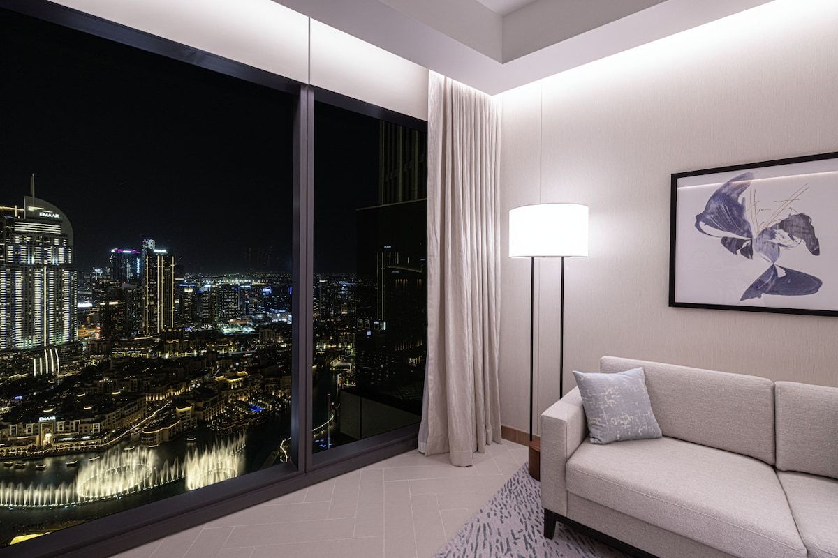 A modern hotel room with a large window offering a city view, a sofa, a floor lamp, and a framed artwork on the wall.