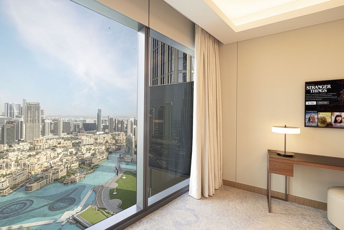 A hotel room with a large window offering a city view, a desk with a lamp, and a poster on the wall.