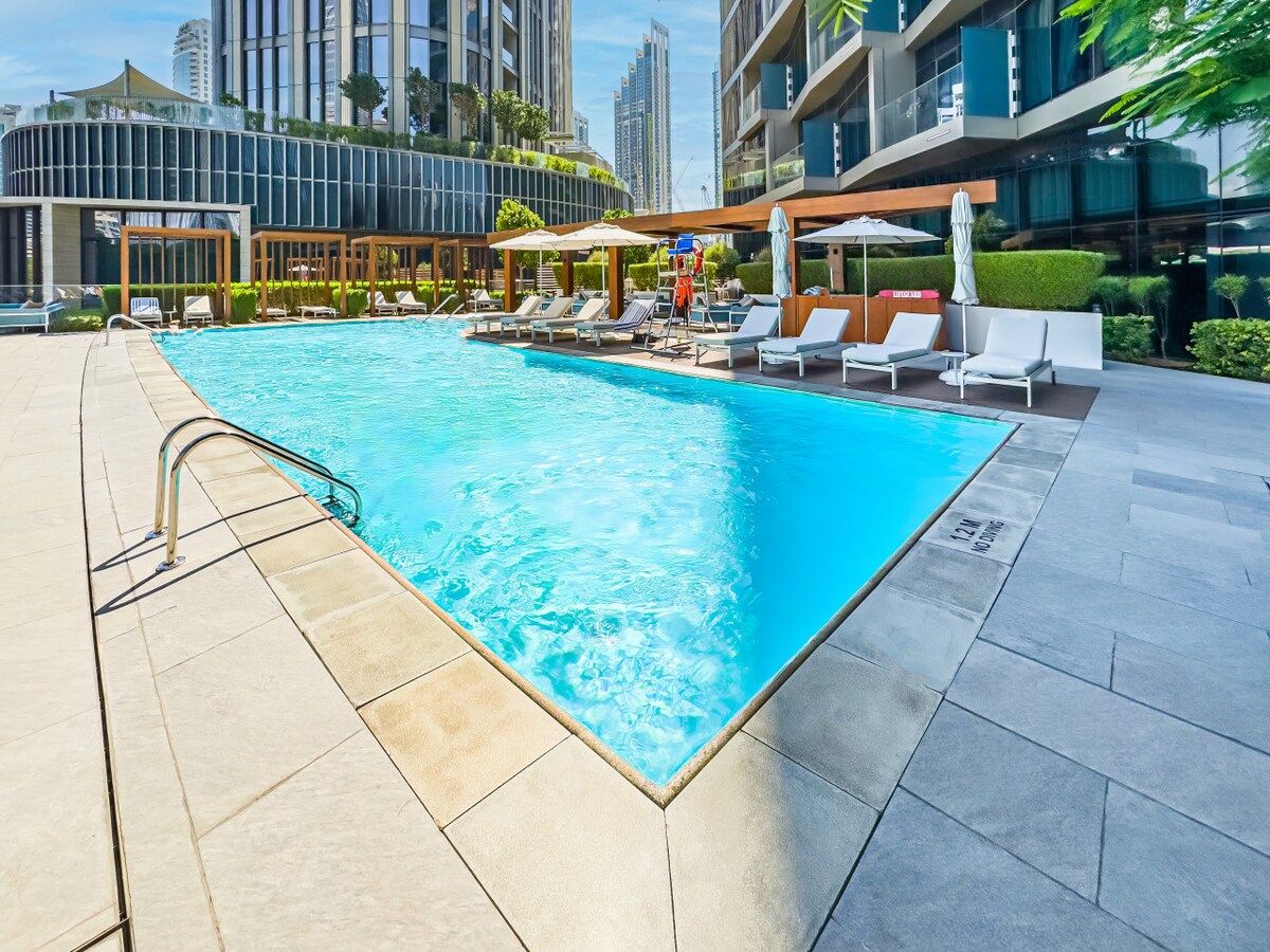 A swimming pool with lounge chairs and umbrellas, surrounded by modern buildings.