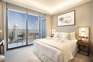A hotel room with a large bed, a desk, a chair, a nightstand, and a view of a cityscape through floor-to-ceiling windows.