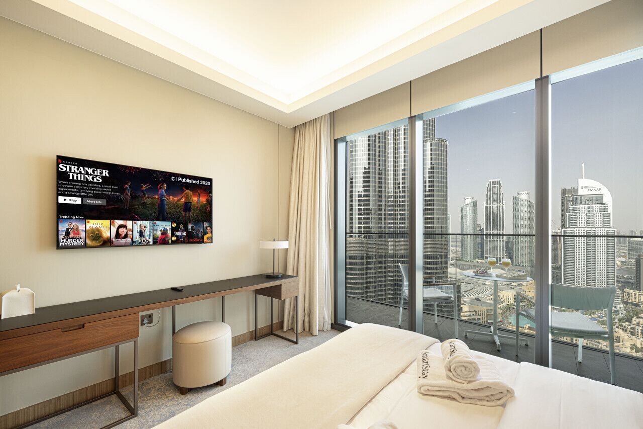 Ein modernes Hotelzimmer mit einem großen Bett, einem Schreibtisch, einem Stuhl und einem Fernseher, der Filmplakate zeigt.