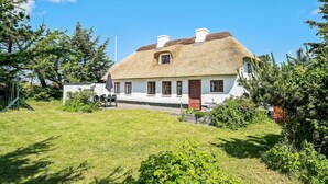 Une maison blanche avec un toit de chaume, une porte rouge et une petite fenêtre. Il y a une pelouse devant et des arbres tout autour.