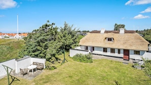 Une maison à toit de chaume, aux murs blancs, entourée d’une pelouse bien entretenue et d’un grand arbre.