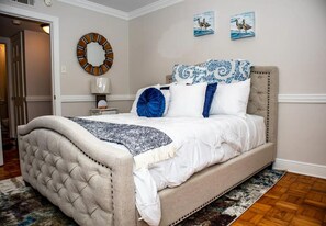 A bedroom with a tufted headboard, a bed with white bedding and blue accents, a round mirror, and two wall-mounted paintings. - 1BR Mobile Condo with Community Pool  (Mobile)