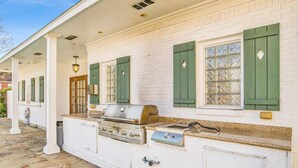 Un bâtiment blanc avec des volets verts, une terrasse pavée de pierres et une cuisinière à gaz.