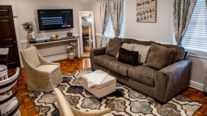 Un salon comprenant un canapé, un pouf et un fauteuil, un téléviseur à écran plat et un ventilateur de plafond.
