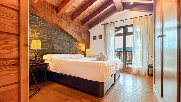 Une chambre avec un plafond en bois, un mur en briques, un lit recouvert de draps blancs, une table de chevet, une lampe et une vue sur l’extérieur par une porte.