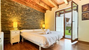 Une chambre avec un mur en pierre, un plafond en bois, un lit avec du linge de lit blanc, deux lampes de chevet et une porte-fenêtre coulissante donnant sur un balcon.