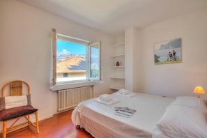 Ein Schlafzimmer mit Bett, Stuhl, Fenster, Heizkörper und einem Regal mit einem Bild.