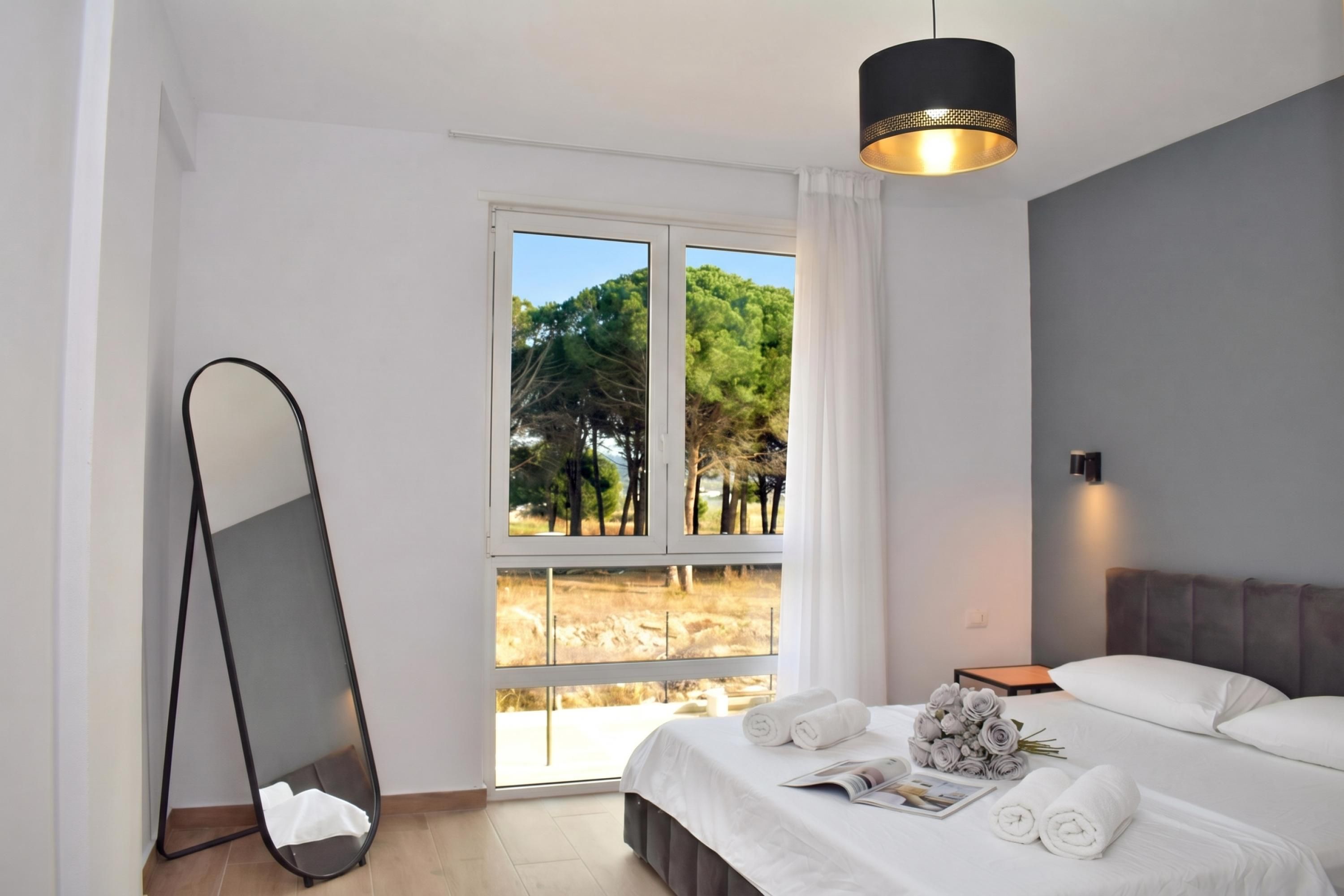 Une chambre moderne avec un grand lit, une table de chevet, une commode, un miroir et une fenêtre donnant sur des arbres.