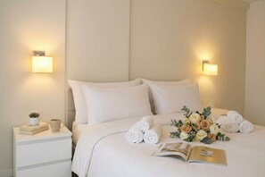 A neatly made bed with rolled towels and a bouquet of flowers, a bedside table with a book and a mug, and wall-mounted lights. - White Moon Apartment 302, Lalzit Bay, Albania (Lalzit Bay)