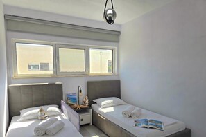 A hotel room with two beds, a nightstand, a lamp, and a window with blinds. - Cozy Apartment in San Pietro Resort, Lalzit Bay, Albania (Lalzit Bay)