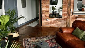 A cozy living room with a brown leather sofa, a patterned rug, and a small ottoman. There are potted plants and a television mounted on a brick wall. - 420 Loft Canada Bridge View - FREE PARKING - FREE WIFI (Detroit)