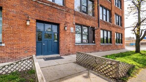 A brick building with a blue door, a small garden area with a wooden fence, and a tree with spring blossoms. - 420 Loft Canada Bridge View - FREE PARKING - FREE WIFI (Detroit)