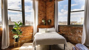 A bedroom with a brick wall, large windows, a bed, a plant, and a view of the city. - 420 Loft Canada Bridge View - FREE PARKING - FREE WIFI (Detroit)