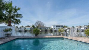 A rectangular swimming pool with clear blue water, surrounded by a white fence, palm trees, and a white building in the background.