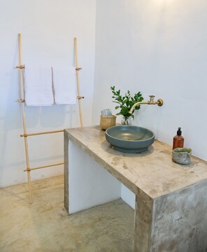 A bathroom with a concrete sink, a bamboo towel rack, a small plant, and a soap dispenser.