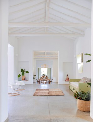 Een ruime woonkamer met een hoge plafond, een eethoek met tafel en stoelen, en een groene bank met een potplant.