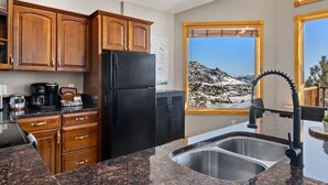 Une cuisine dotée de plans de travail en granit, d’un réfrigérateur noir, de meubles en bois et d’une fenêtre offrant une vue sur les montagnes.
