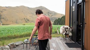 Une personne se tient sur une terrasse en bois, un ordinateur portable à la main, et contemple un champ verdoyant et des montagnes.