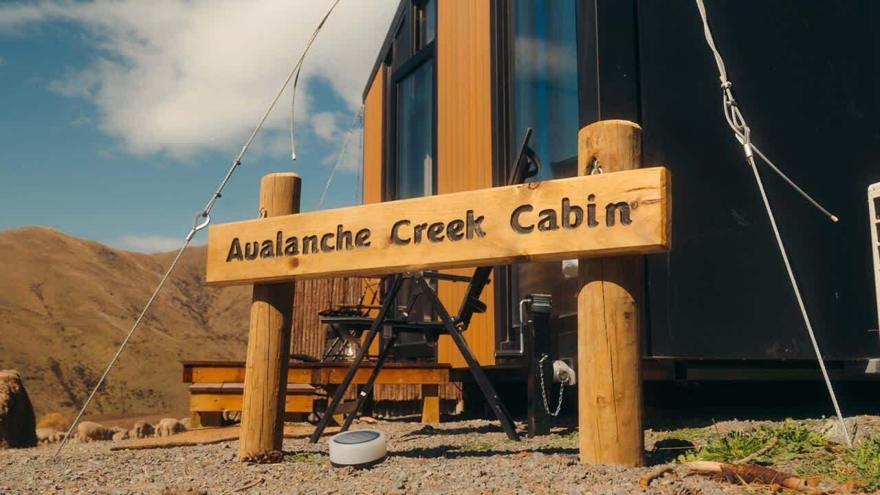 A wooden sign for 'Avalanche Creek Cabin' with a rustic outdoor setting, including a wooden bench and a dog bowl.