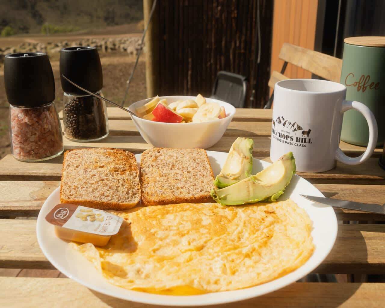 Een ontbijtbord met toast, avocado en roerei, een schaal fruit, en een koffiemok op een houten tafel.