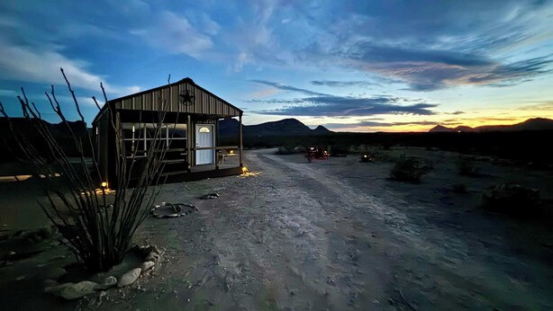 A rustic wooden building with a door and windows, situated on a dirt road with sparse vegetation. - Desert Quail Oasis: Style, Comfort & Easy Access (Terlingua)