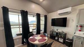 A dining area with a round table, black chairs, a flat-screen TV mounted on the wall, and a view of the outdoors through large windows. - Desert Quail Oasis: Style, Comfort & Easy Access (Terlingua)