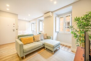 A modern living room with a light-colored sectional sofa, a wooden floor, and a large window with a view. - GuestReady - Charming Retreat in Historic Lisbon (Lisbon)