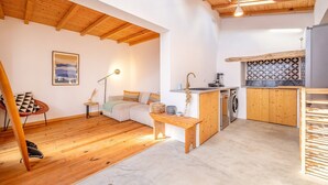 Una sala acogedora con un sofá, un banco de madera, una lámpara de pie y una mesa de comedor. Hay una zona de cocina con un fregadero, estufa y lavadora. El cuarto tiene suelo de madera y techo inclinado con vigas de madera.