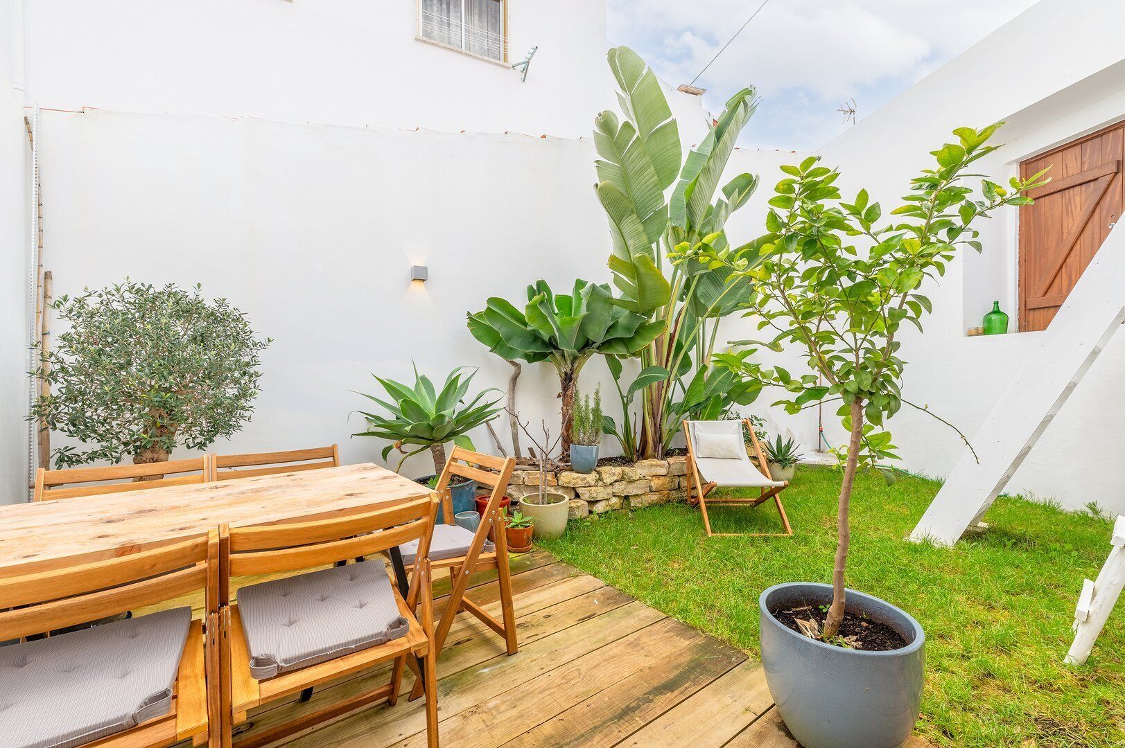 Una terraza de madera con una mesa y sillas, rodeada de macetas con plantas y un árbol pequeño.
