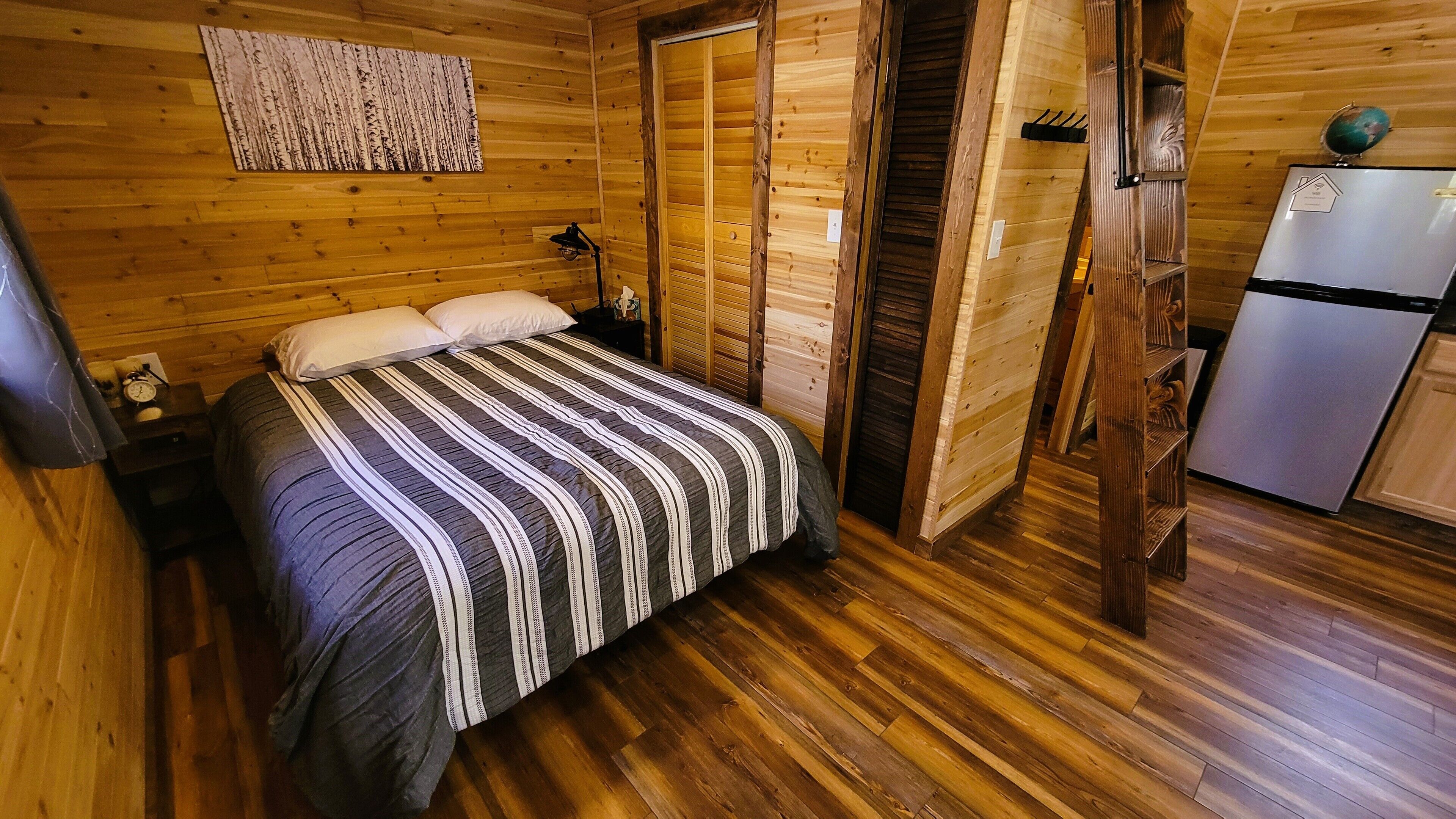A wooden cabin bedroom with a bed, a nightstand, a globe, and a refrigerator.